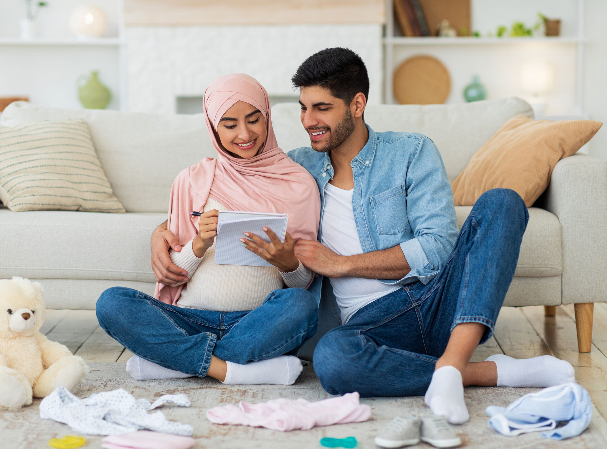 A couple with a pregnant woman sit on the floor around baby clothes and toys.
